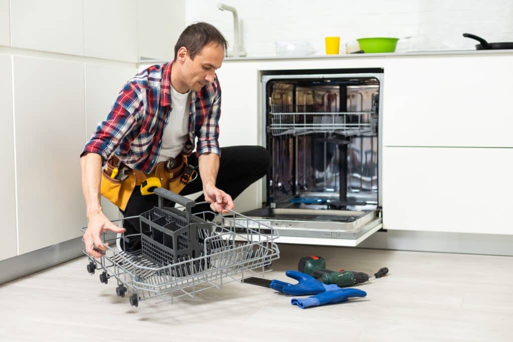 Plumber repairing an open dishwasher while removing the lower dish rack in a modern kitchen.