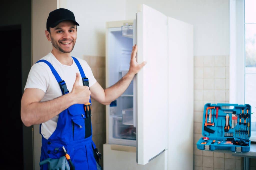 HVAC technician standing beside an open refrigerator, giving a thumbs-up with a tool kit nearby.