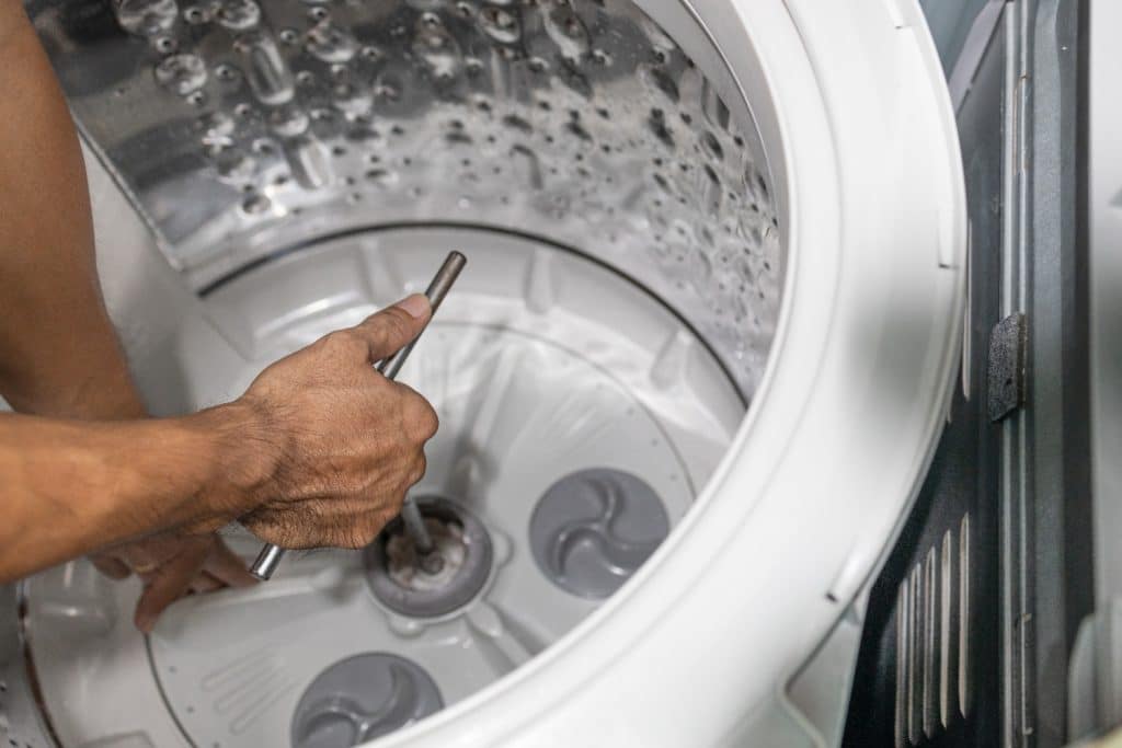 A technician repairing the inside of a front-loading washing machine, using a metal tool to work on the drum and internal components.